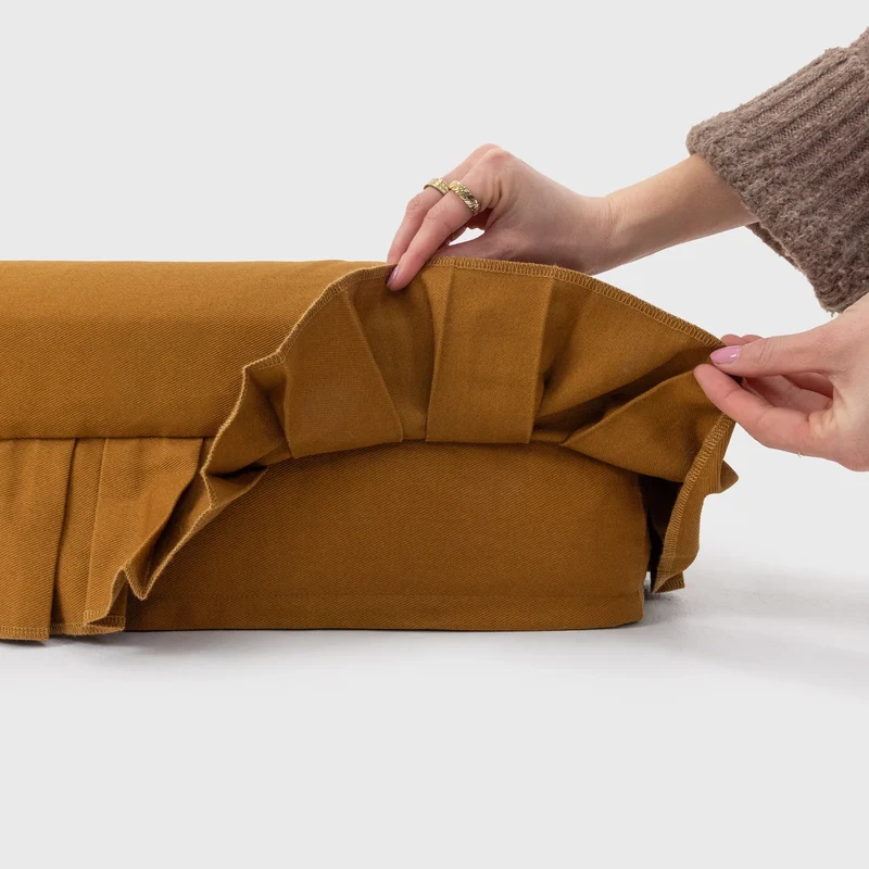 Nostalgia Cover in Toffee: hands lifting the pleated skirt to show the plain toffee side of the bed underneath.