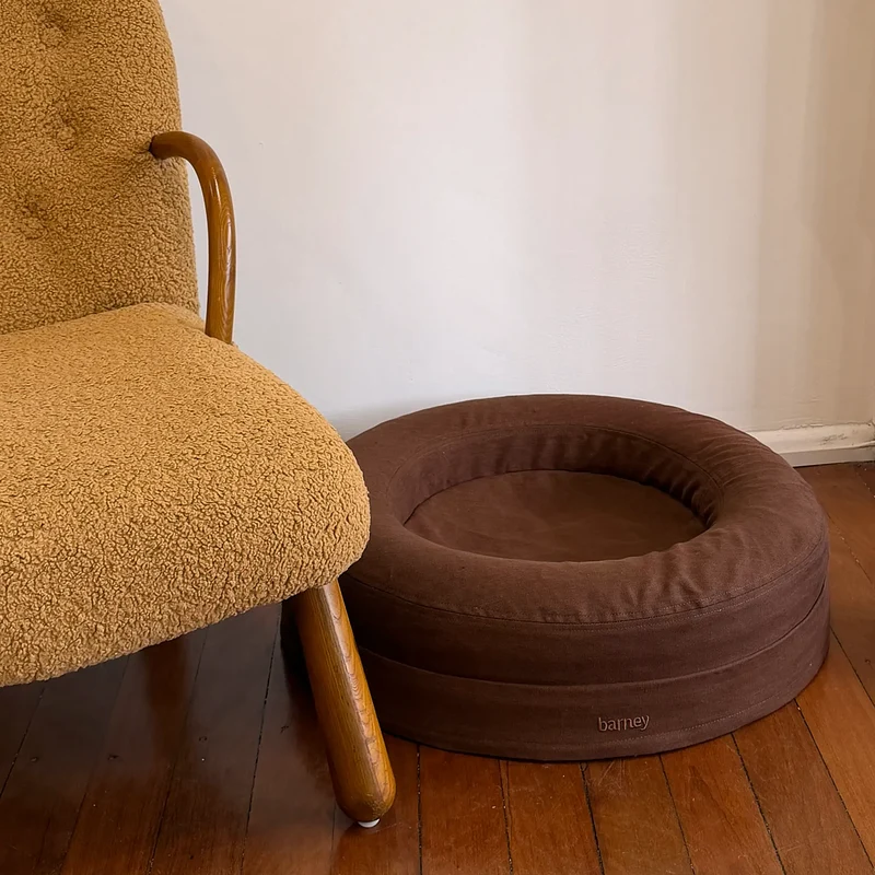 Linen Cover Curl in Moose: the empty round chocolate brown linen bed on a timber floor beside an armchair.