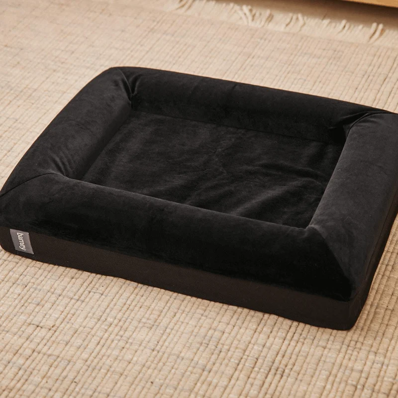 Original Cover in Jet Black: an empty bed on a woven rug, with soft black velvet bolsters on all four sides.