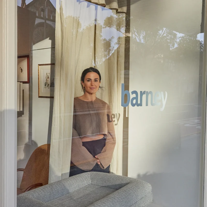Founder Julia Sakr standing inside the Barney Boutique window behind "barney" on the glass, a gray herringbone bed in front.